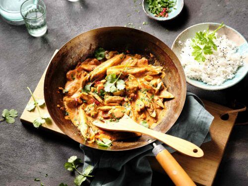 Schweine-Curry mit Chicorée, serviert in einer dekorativen Pfanne, umgeben von frischen Kräutern und Gewürzen.