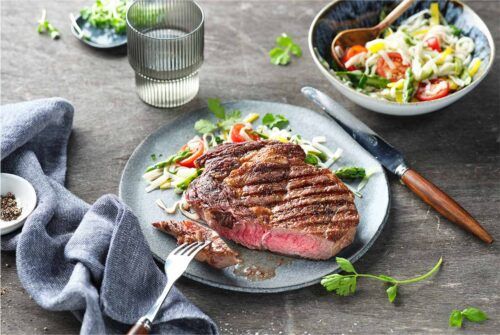 Gegrillte Rib Eye Steaks serviert mit Glasnudelsalat, Koriander und frischem Thai-Basilikum auf einem Teller.